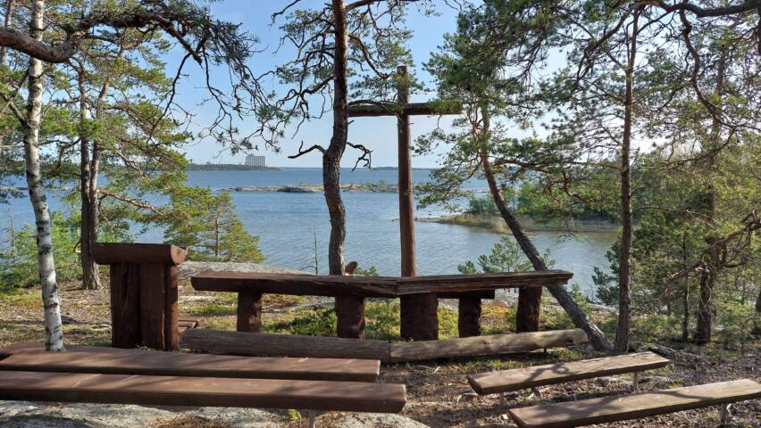 Puiset penkit kalliolla, takana puinen risti ja taustalla merinäköala.