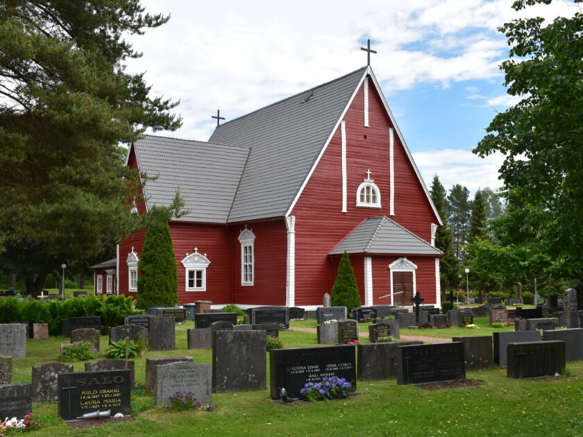 Etualalla hautakiviä, taustalla punainen puukirkko.