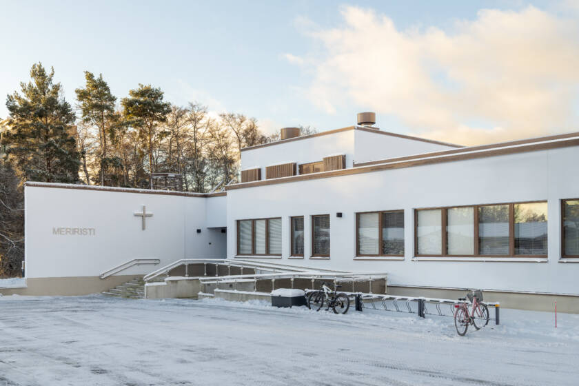 Valkoinen rakennus, etualalla parkkipaikka, taustalla puita ja taivasta.