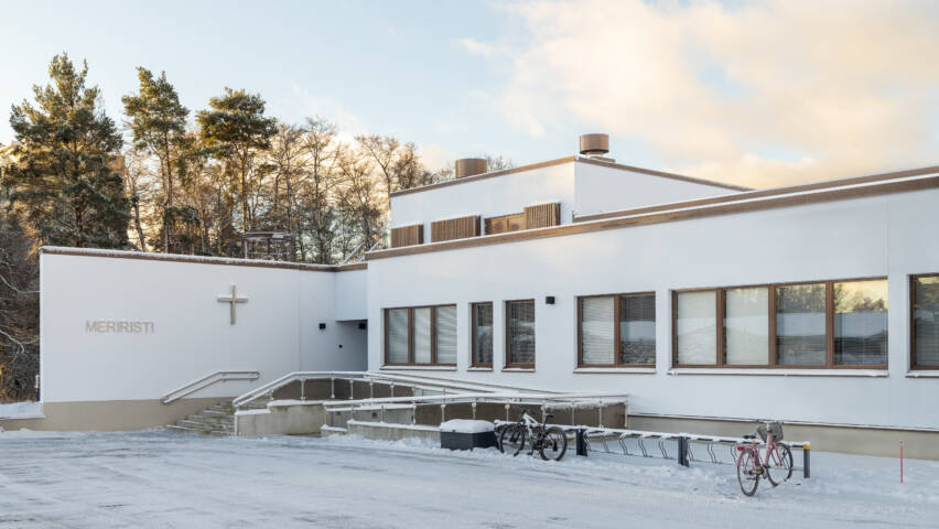 Valkoinen rakennus, etualalla parkkipaikka, taustalla puita ja taivasta.