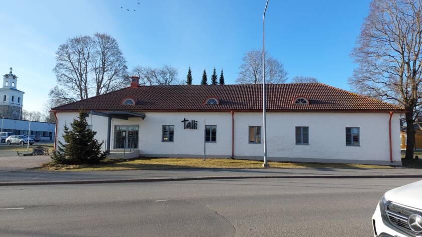 Pitkä valkoinen rakennus, taustalla puita ja kirkko.