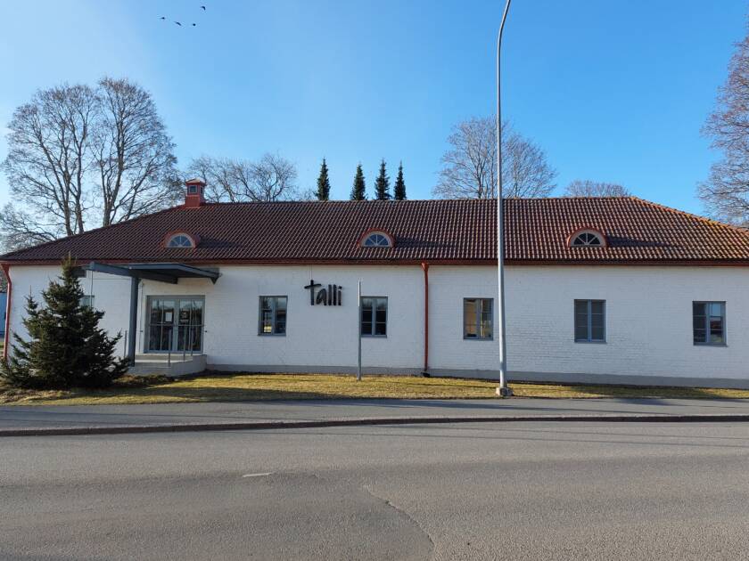 Pitkä valkoinen rakennus, taustalla puita ja kirkko.