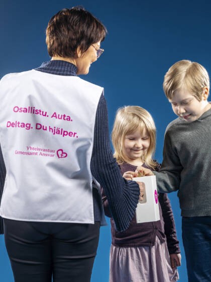 Kaksi lasta pudottavat kolikon keräyslippaaseen, joka on naisen kädessä. Nainen on selin kameraan.