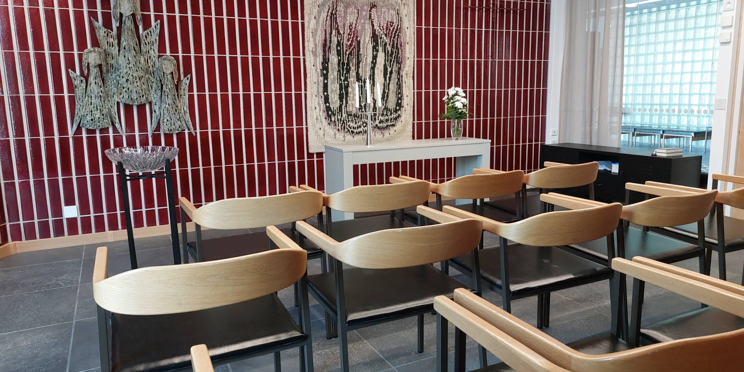 Tuoleja rivissä. Punainen kaakeliseinä, jossa ryijy. Kastemalja.
