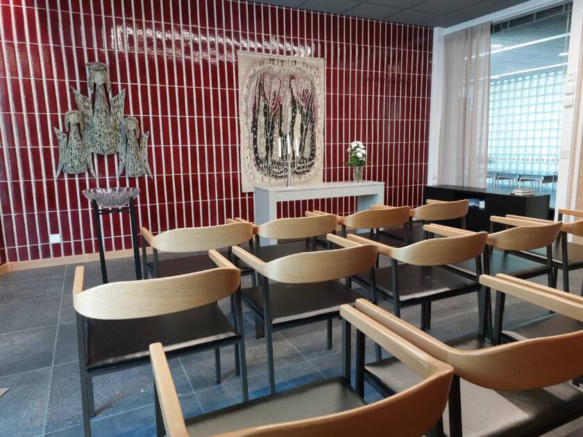 Tuoleja rivissä. Punainen kaakeliseinä, jossa ryijy. Kastemalja.