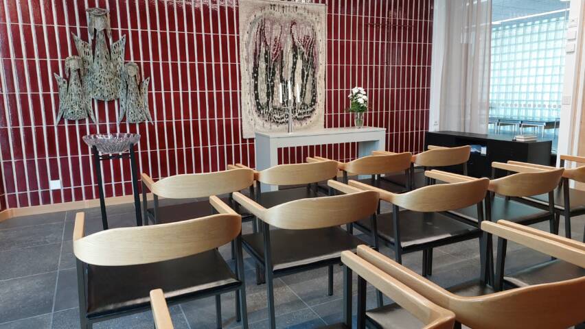 Tuoleja rivissä. Punainen kaakeliseinä, jossa ryijy. Kastemalja.