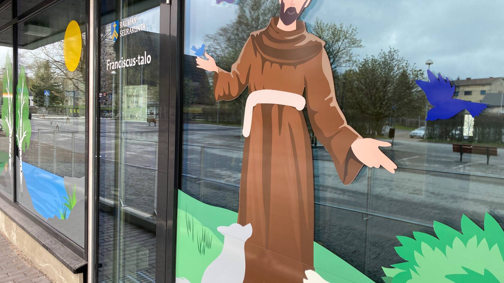 Oven vieressä ikkunateippaus Franciscuksesta, jolla ruskea kaapu ja jalkojen juuressa eläimiä.