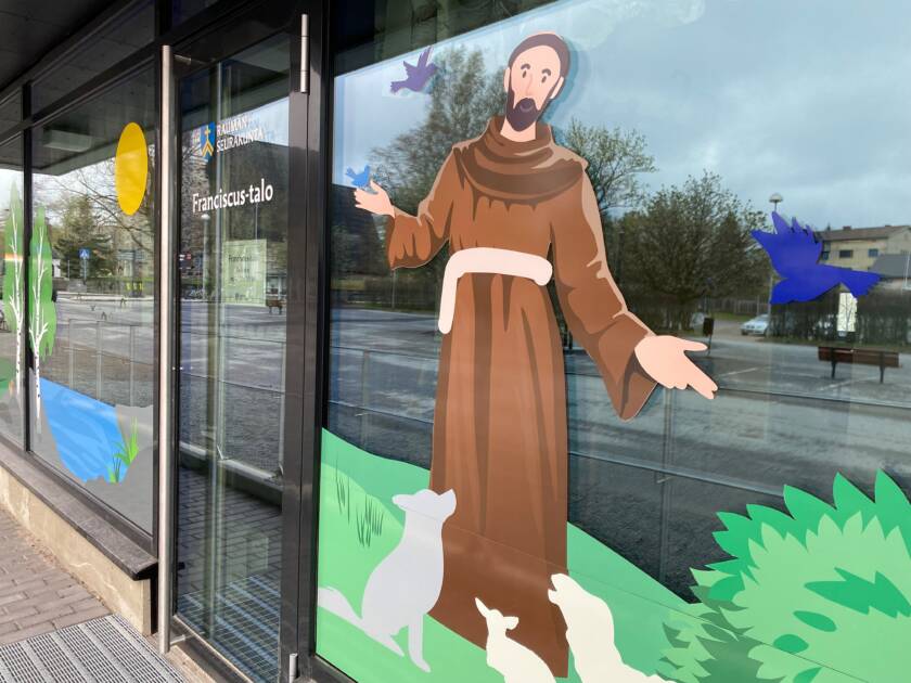 Oven vieressä ikkunateippaus Franciscuksesta, jolla ruskea kaapu ja jalkojen juuressa eläimiä.