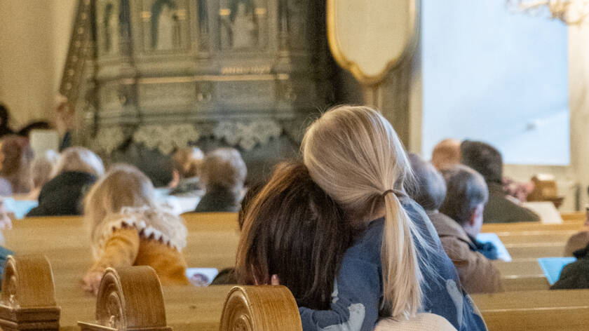 Lapsi halaa äitiään kirkon penkissä, taustalla Pyhän Ristin kirkon saarnastuoli ja muita perhekirkossa kävijöitä.