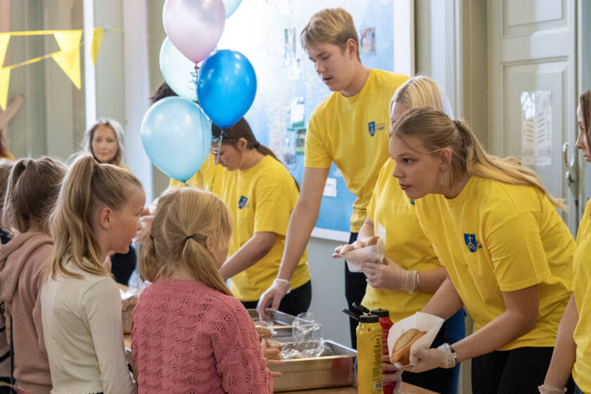 Lapset jonottavat hodareita. Ruokaa jakavat keltapaitaisia nuoria. Taustalla ilmapalloja.