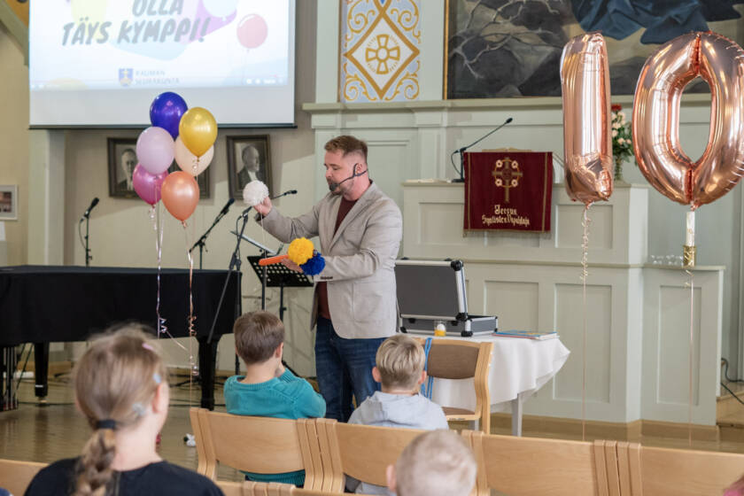 Etualalla lapsia, taustalla ilmapallot, joista muodostuu numero kymmenen. Taustalla taikuri.