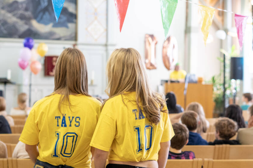 Kaksi nuorta keltaisissa paidoissa, joiden selässä lukee: Täys 10. Taustalla ilmapalloja ja lapsia.