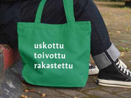 Vihreä kangaskassi, jossa on valkoiset sanat "uskottu", "toivottu" ja "rakastettu". Taustalla henkilö istuu kivetyksellä.