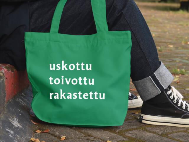 Vihreä kangaskassi, jossa on valkoiset sanat "uskottu", "toivottu" ja "rakastettu". Taustalla henkilö istuu kivetyksellä.