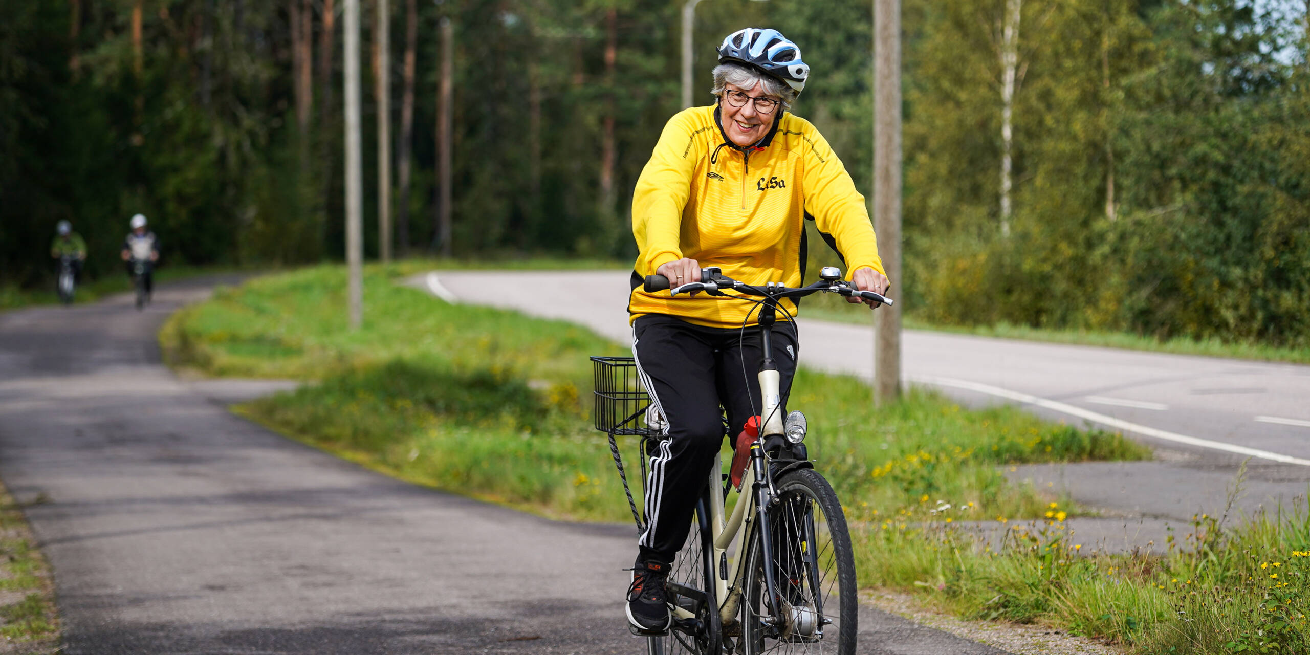 Nainen keltaisessa takissa pyöräilee. Taustalla tie ja metsää.