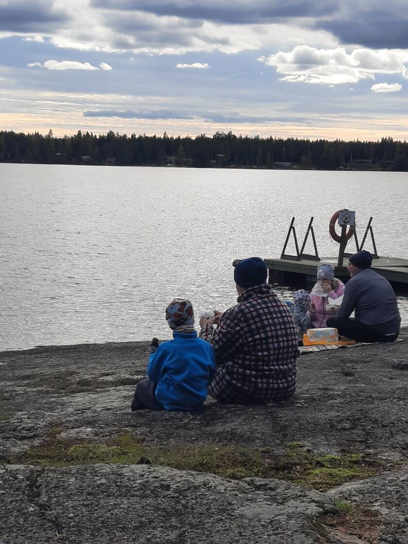Ihmisiä istuu rantakalliolla, taustalla järvimaisema ja laituri.