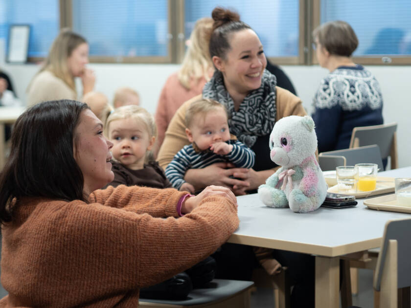 Nainen nojaa pöytää vasten, jonka ääressä istuu nainen lasten kanssa. Pöydällä pehmolelu ja tarjottimia.
