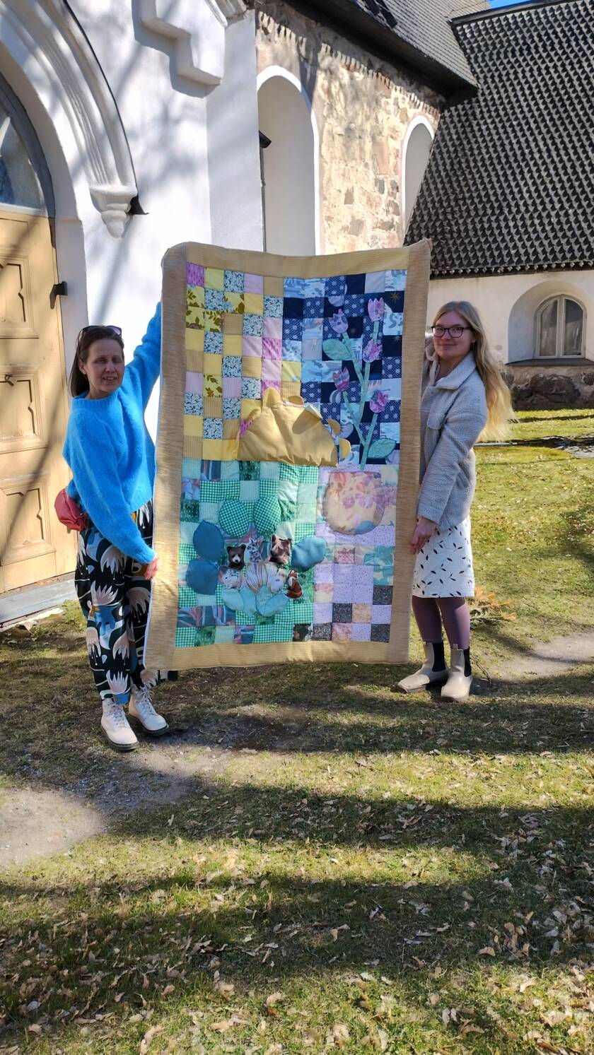 Kaksi naista pitelee isoa tilkkutäkkiä kirkon pihalla.