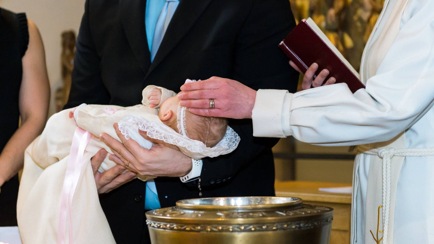 Kuvassa pieni vauva kasteella Pyhän Ristin kirkossa. Kuvassa näkyy kastemalja, papin käsi kastamassa. Vauva on kummin sylissä.